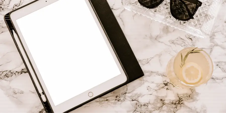 Tablet with stylus on marble surface next to drink glass and sunglasses placed nearby