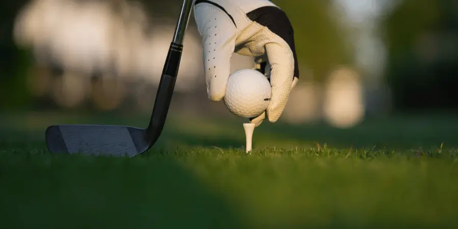 Hand placing golf ball on tee with golf club nearby on grass, outdoor sports scene with shallow depth of field
