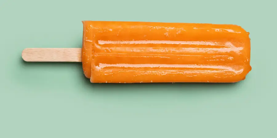 Single orange popsicle on a stick placed horizontally on a green background surface